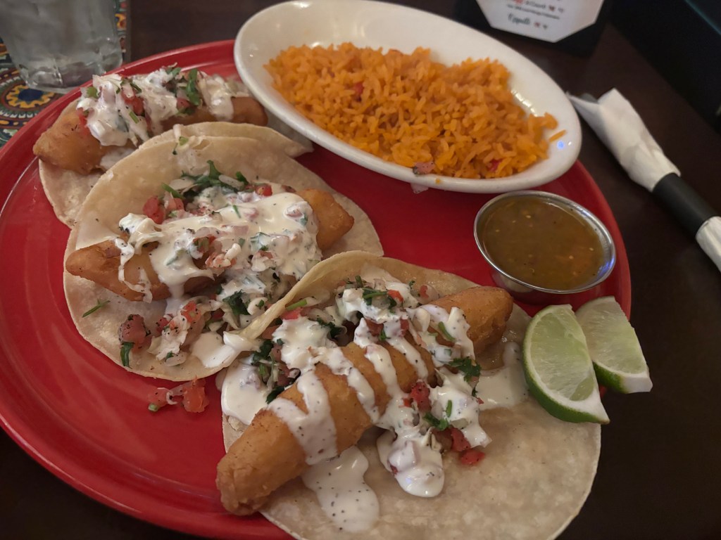 Three tacos filled with crispy fried fish, topped with creamy sauce, diced tomatoes, and cilantro, served on corn tortillas with lime wedges and a side of orange rice and salsa.