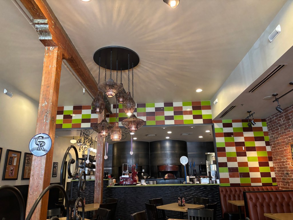 Interior view of a restaurant featuring exposed wooden beams, colorful checkered tiles, and pendant lighting. A pizza oven is visible in the background along with seating areas.