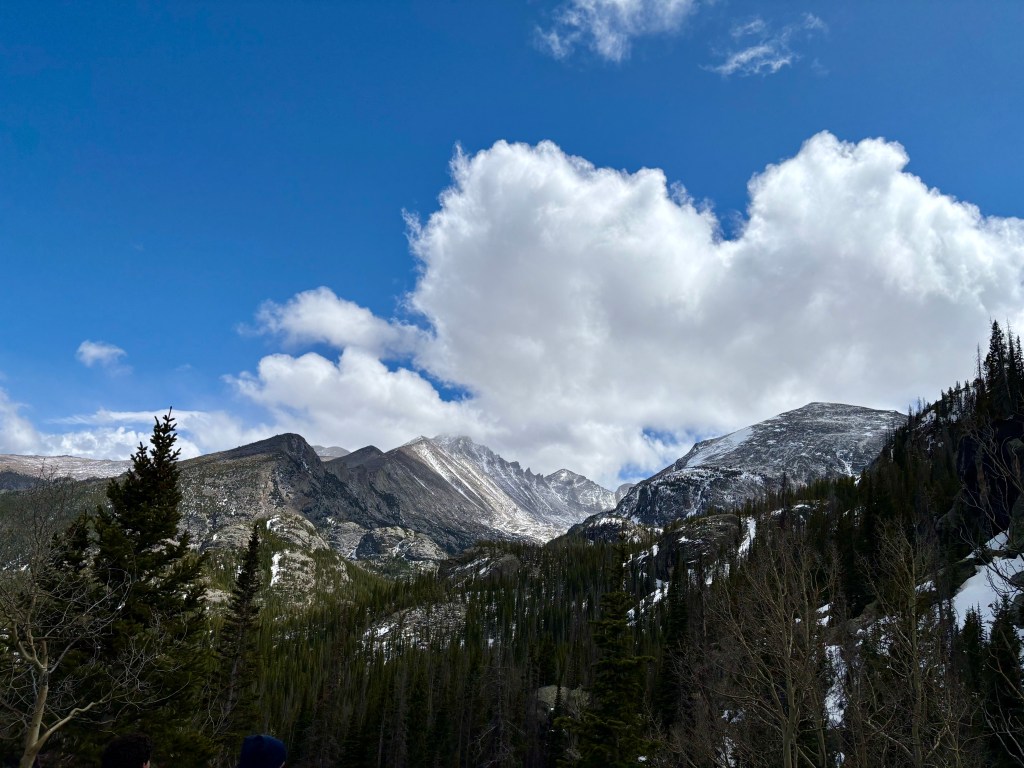 Snow-capped mountains under a blue sky with fluffy white clouds, surrounded by evergreen trees.