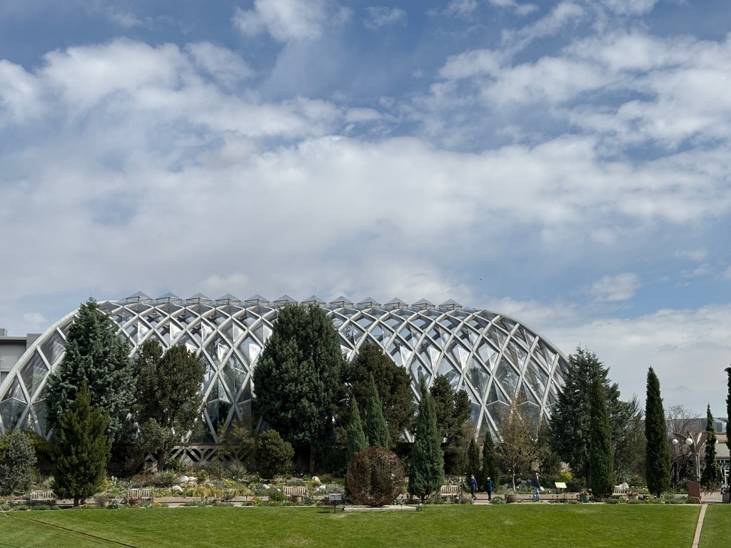 A large glass dome structure surrounded by trees and plants, with a blue sky and clouds in the background.