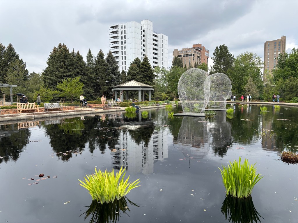 A serene garden scene featuring a still pond reflecting trees and buildings, with wire sculptures in the water, surrounded by visitors and benches.