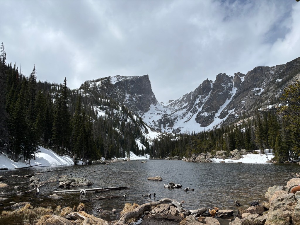 A serene mountain lake surrounded by snow-capped peaks and pine trees under a cloudy sky.