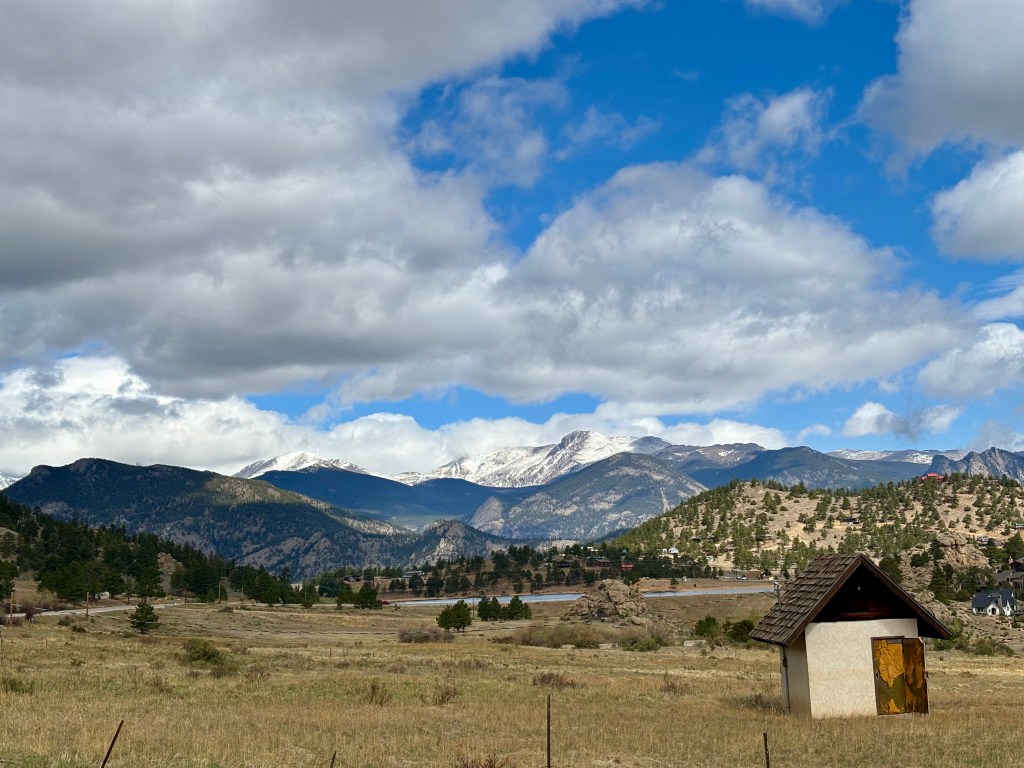 A scenic view of mountains under a partly cloudy sky, with a small structure in the foreground and a grassy plain.