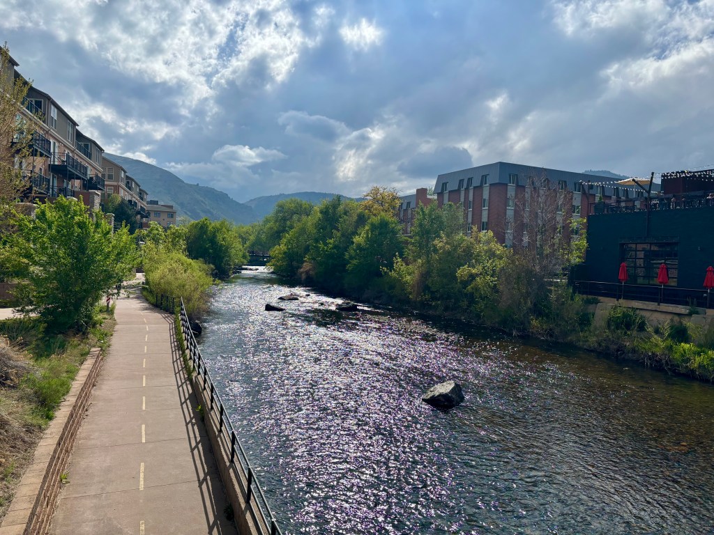 A scenic view of a river with a pathway alongside, surrounded by greenery and modern buildings under a partly cloudy sky.