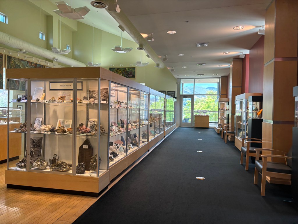 Interior view of a mineral museum display area showcasing glass cases filled with various minerals and gemstones, with seating areas and large windows letting in natural light.