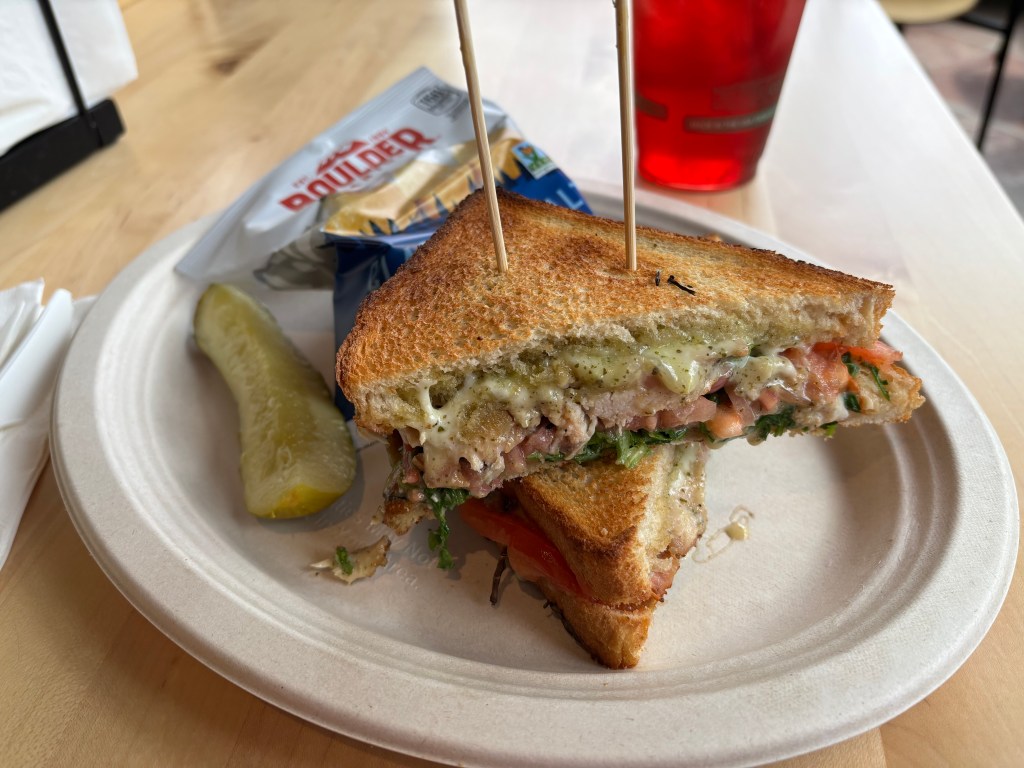 A close-up of a toasted sandwich filled with lettuce, tomato, and meat, partially sliced and presented on a paper plate, accompanied by a pickle and a bag of chips.