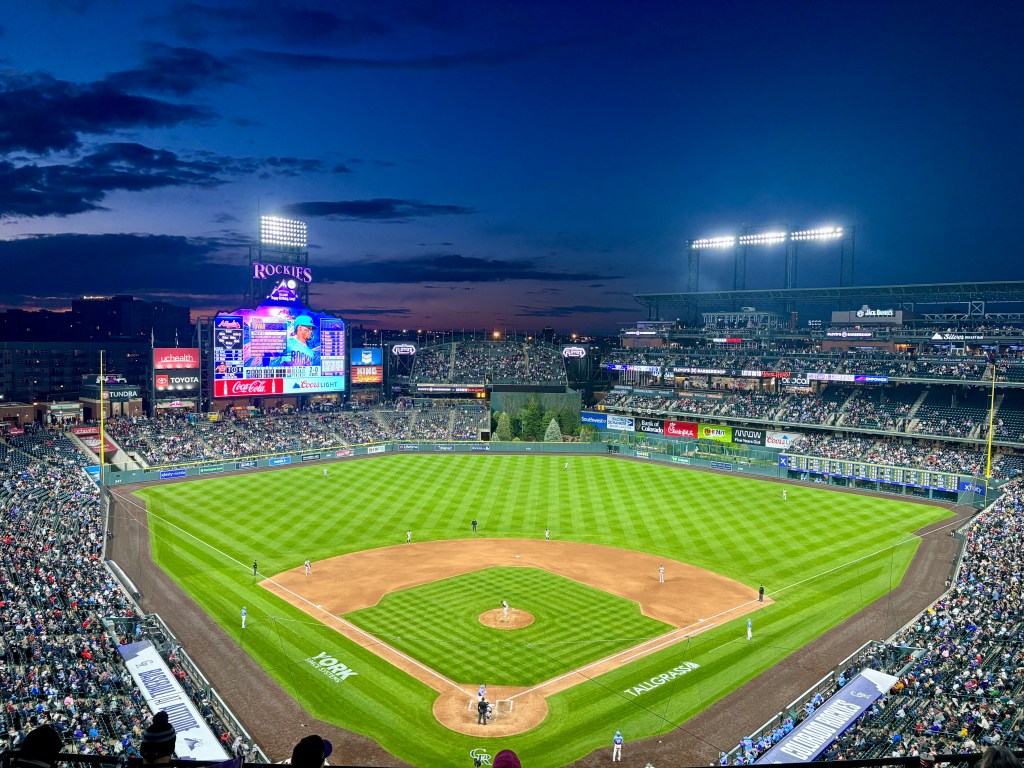 Aerial view of a baseball stadium during twilight, showcasing a lively crowd, a vibrant scoreboard displaying team information, and a well-manicured field.