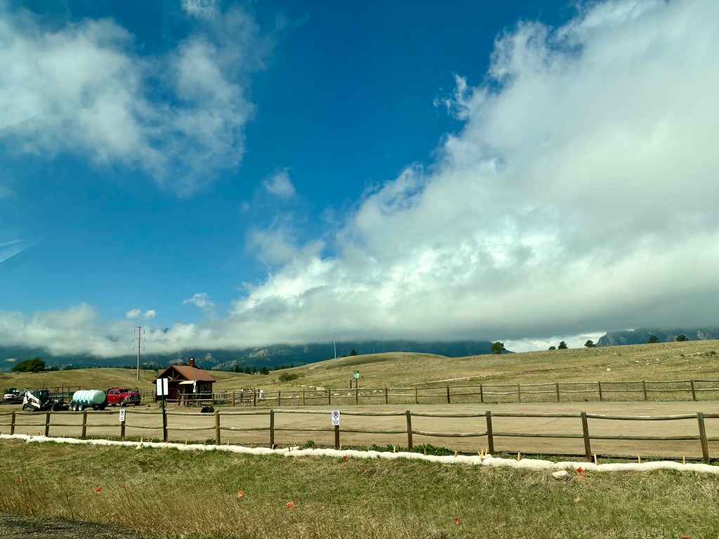 A scenic landscape featuring rolling hills under a blue sky with scattered clouds. In the foreground, a wooden fence encloses a dirt area with a small building and vehicles, while mountains loom in the background.