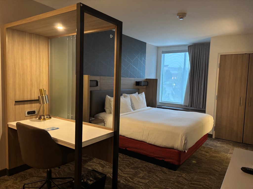 Modern hotel room featuring a queen bed, a work desk with a lamp, and a large window with curtains.