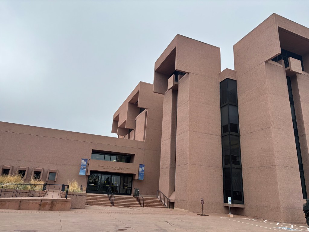 Exterior view of a modern architectural building with angular features and a textured facade, featuring large glass windows and entry steps.