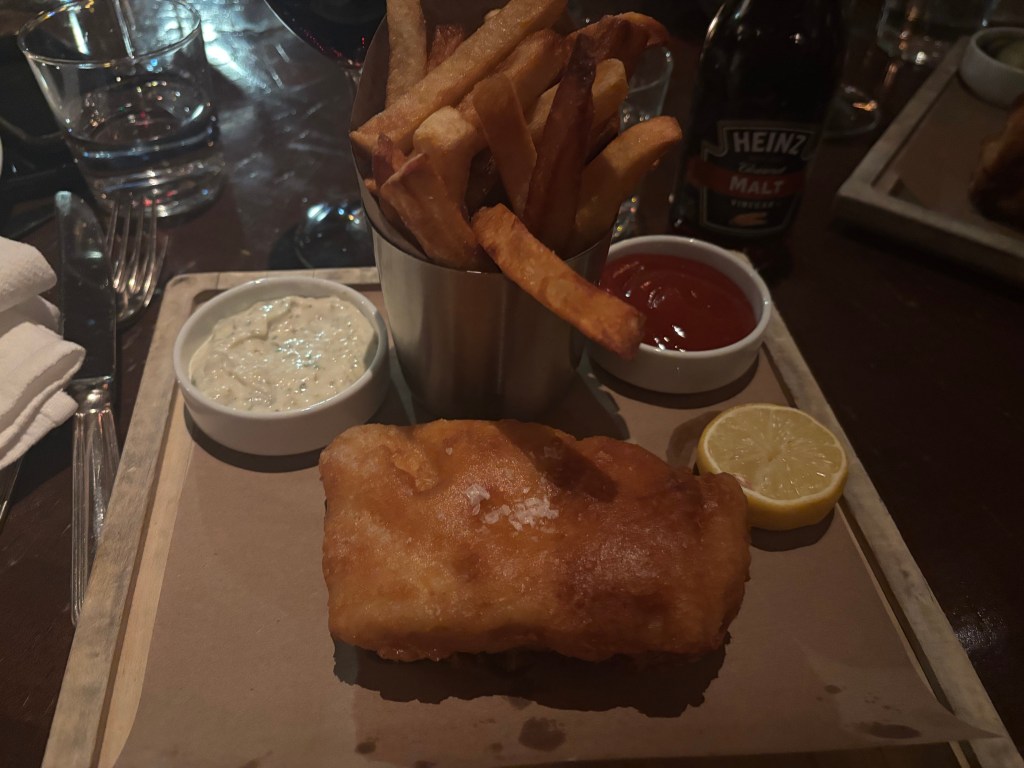 A plate featuring a piece of fried fish, accompanied by a serving of French fries in a metal cup, tartar sauce in a small dish, ketchup in another small dish, and a lemon wedge.